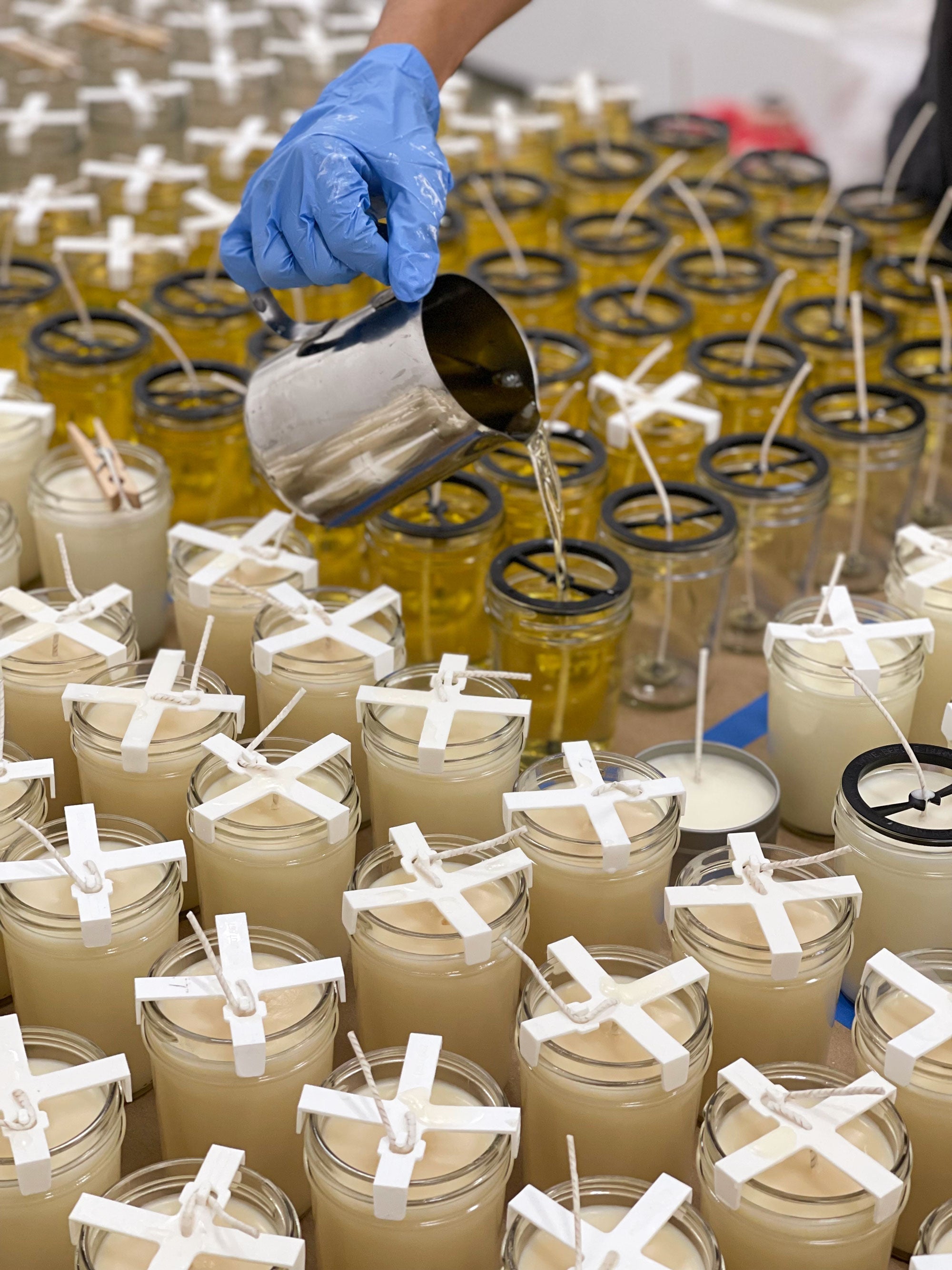 a gloved hand pouring liquid soy wax into rows of candle jars