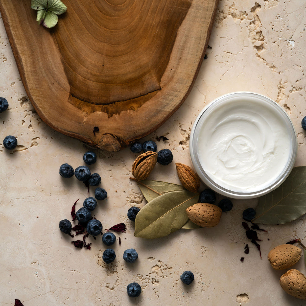 White container of cream on a rustic surface with blueberries, almonds, and a wooden board.