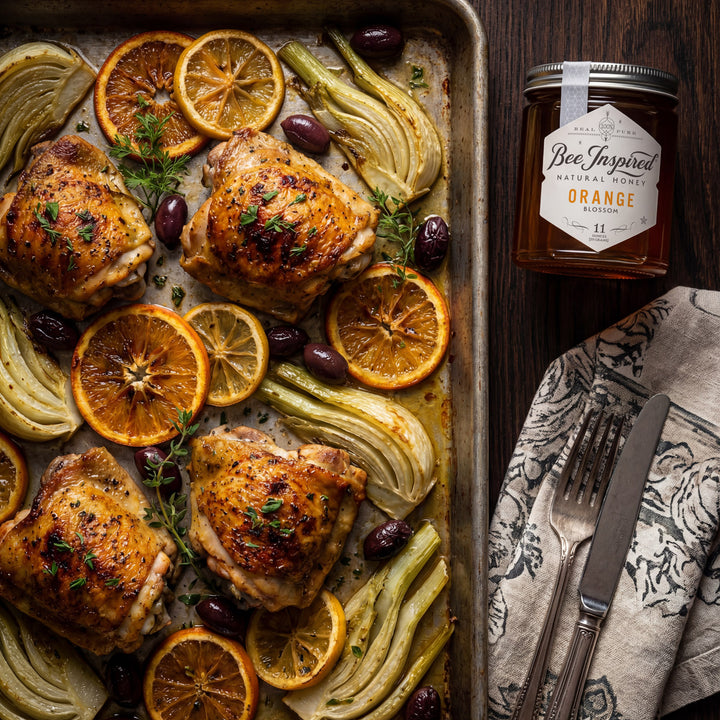 Roasted chicken with oranges and herbs on a baking tray, accompanied by a jar of honey.
