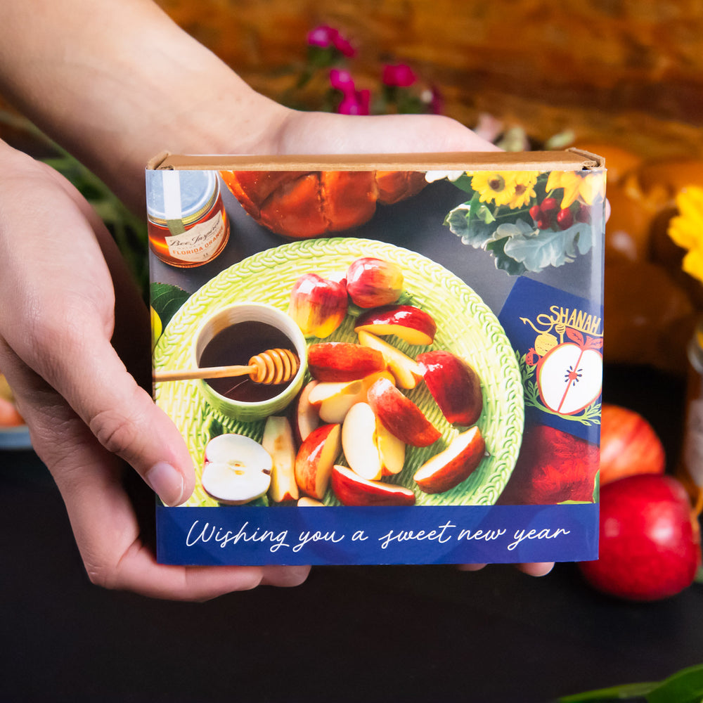 Hand holding a box with apples, honey, and a jar on a colorful background