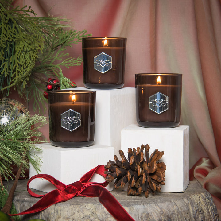 Three lit candles with a geometric logo on brown glass containers, surrounded by greenery and a pine cone on a wooden surface.