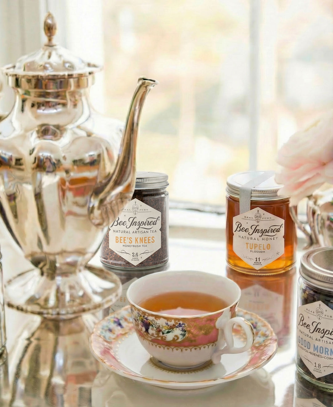 Tea cup with floral design on a saucer, silver teapot, and jars of 'Bee Inspired' honey on a reflective surface.