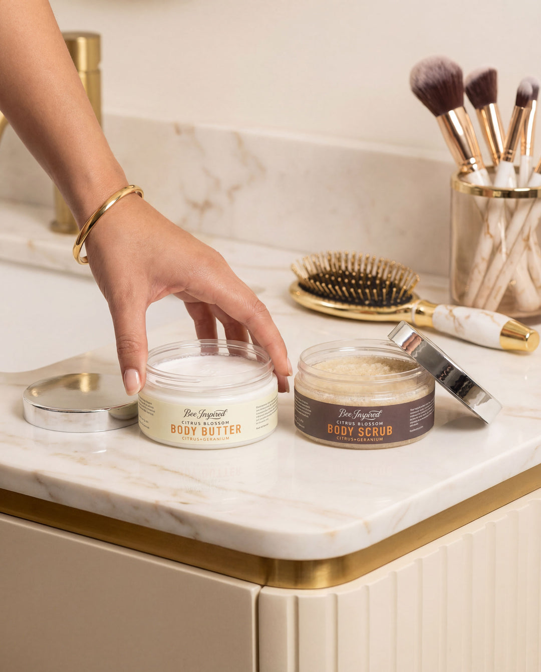 Hand reaching for a jar of body scrub on a marble countertop with makeup brushes in the background.