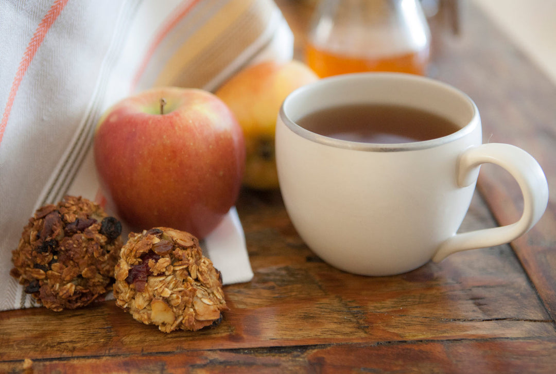White mug with hot beverage, apple, and cookies on a wooden surface