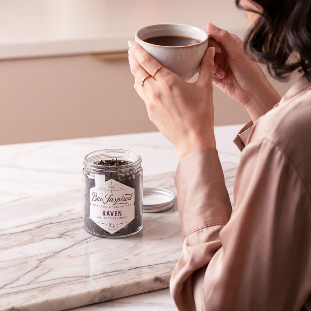 Person holding a cup of raven tea with a tea jar labeled 'Bee Inspired' on a marble surface.