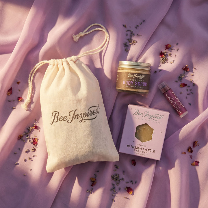 Bee Inspired skincare products including a drawstring bag, body scrub, bar soap, and lip balm on a pink fabric background.