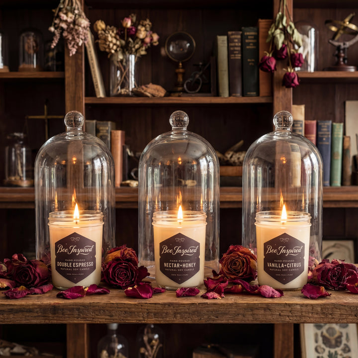 Three lit candles in glass domes on a wooden surface with flowers and books in the background