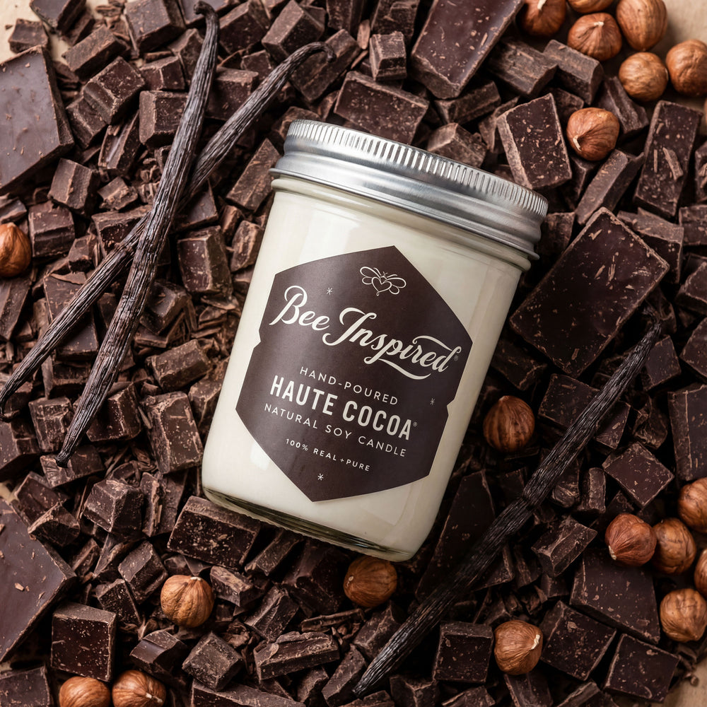Candle jar labeled 'Bee Inspired' surrounded by chocolate pieces and vanilla beans.