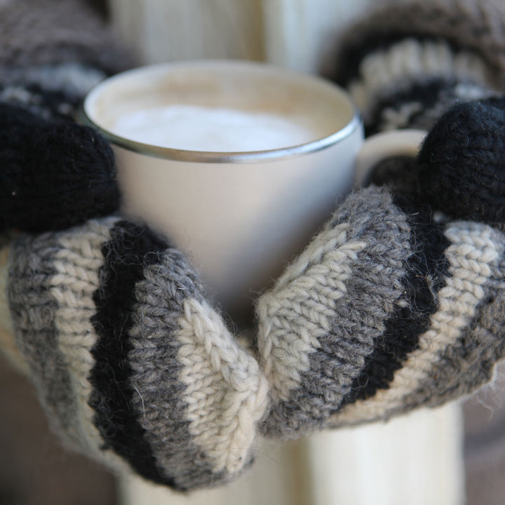 Pair of hands wearing striped mittens holding a cup of coffee.