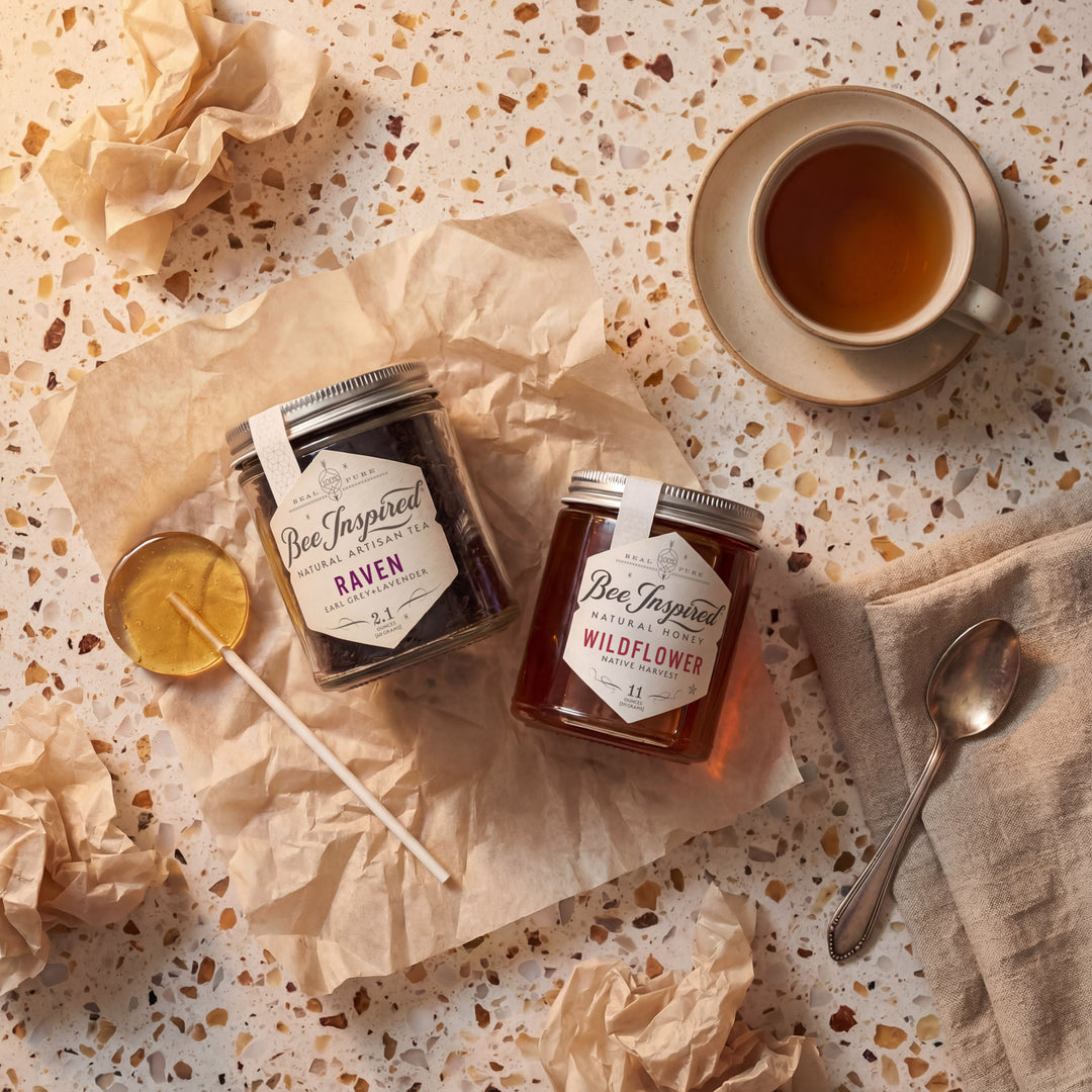 A jar of Raven Tea and a jar of Wildflower honey on a textured surface with a cup of tea and a spoon.