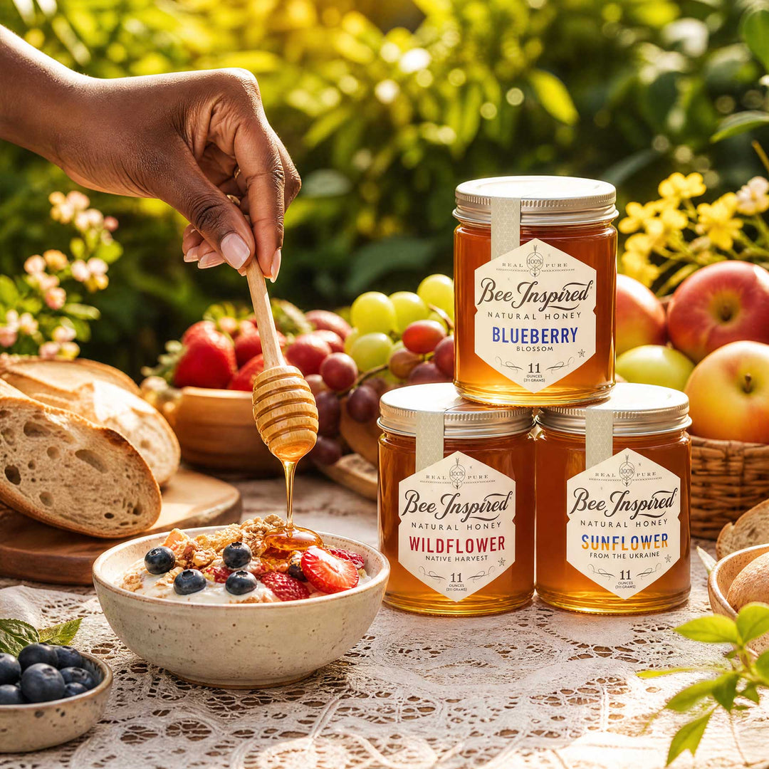 Three glass jars of Bee Inspired Honey arranged on a table outdoors, with a hand holding a honey dipper over a bowl of oatmeal with berries. Bread, grapes, apples, and greenery are visible in the background.