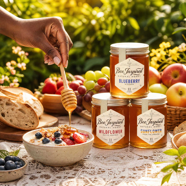 Three glass jars of Bee Inspired Honey arranged on a table outdoors, with a hand holding a honey dipper over a bowl of oatmeal with berries. Bread, grapes, apples, and greenery are visible in the background.