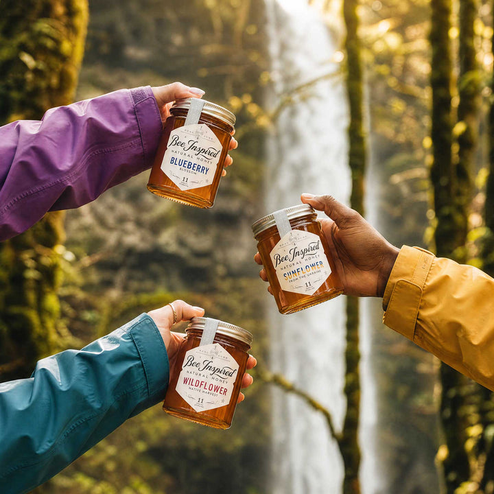 Three people in outdoor rain jackets hold jars of Ben Joyasut honey in a forest setting with a waterfall in the background. The jars are labeled Blueberry, Honeycomb, and Wildflower.