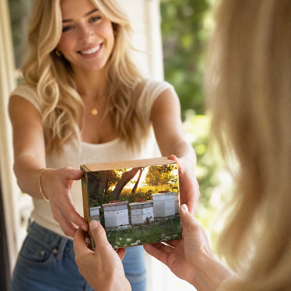 A woman smiling and holding out a small printed box with a photo of beehives, while another person in the foreground holds it. The scene is softly lit and appears to be a lifestyle shot.