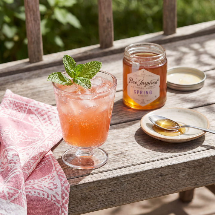 Glass of strawberry honey lemonaide iced drink with a mint leaf on a wooden table, next to a jar labeled &