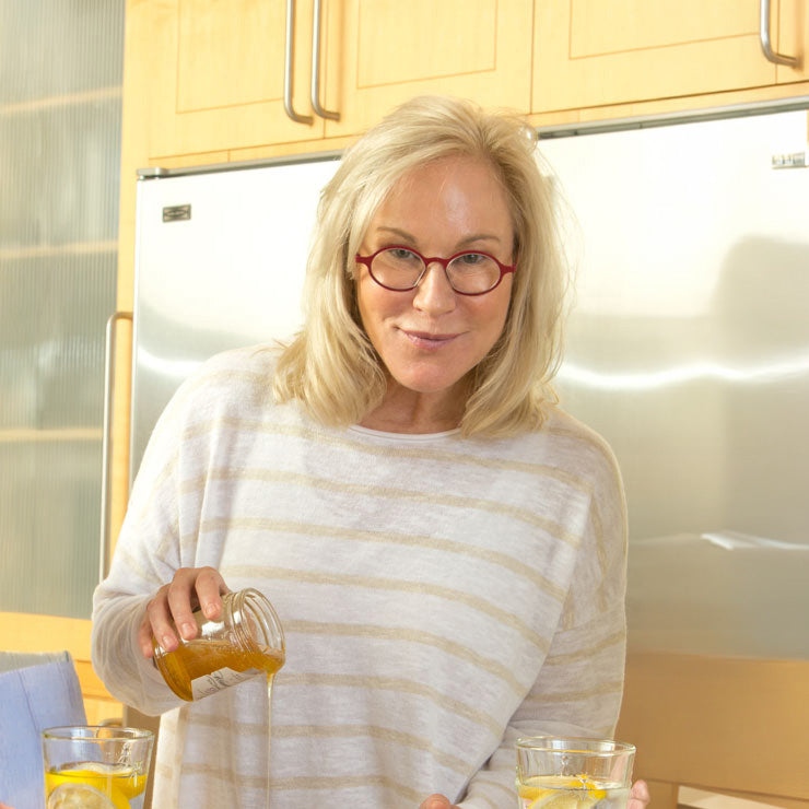 A woman with blonde hair and red-rimmed glasses wearing a white and beige striped long-sleeve shirt, pouring honey from a glass jar into a glass of water in a kitchen setting.