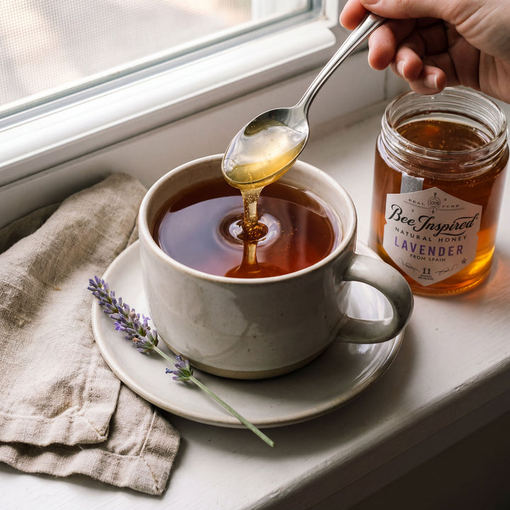 Honey being drizzled into a cup of tea with a jar of lavender honey on a windowsill.