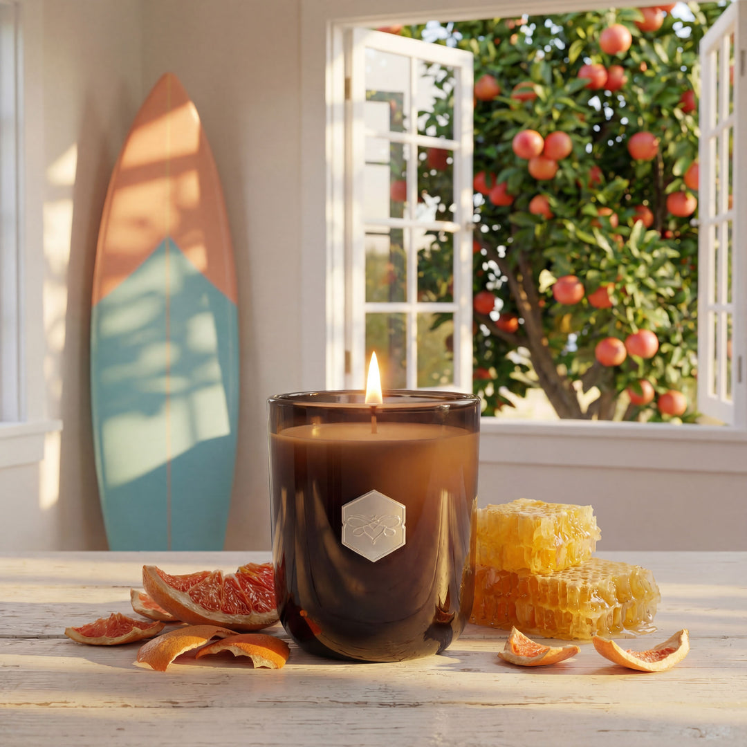 Candle in a dark glass holder with a hexagonal logo on a wooden surface, with dried fruit and a surfboard in the background.