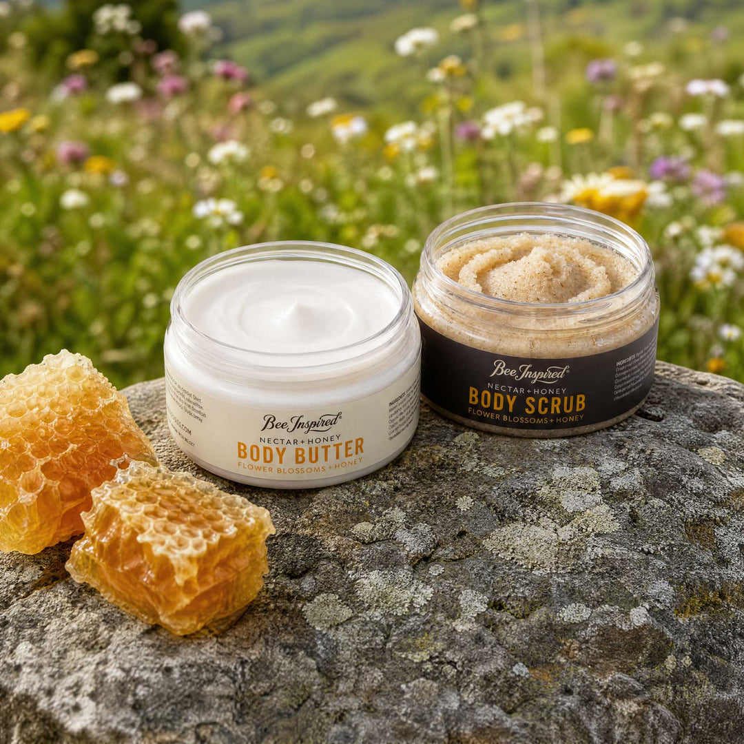 Two jars of Bee Inspirational body skincare products, Body Butter and Body Scrub, displayed on a stone with honeycomb in a blurred meadow background.