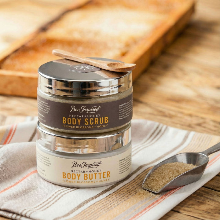 Two containers of Bee Inspired Nectar Honey Body Scrub and Body Butter on a wooden surface with a wooden spoon.