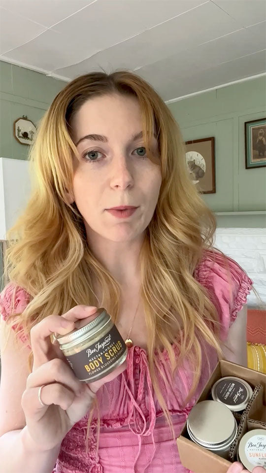 Woman holding a jar of body scrub in a room with framed pictures on the wall.