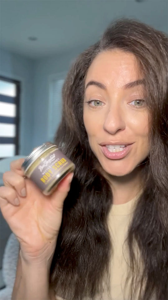 Woman holding a jar of body scrub in a room with light blue walls.