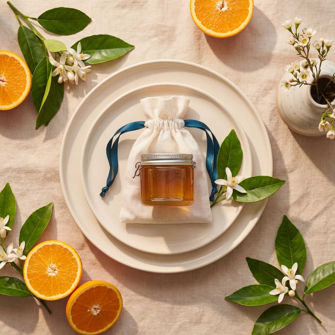 Small jar of honey in a drawstring bag on a white plate with oranges and flowers around.