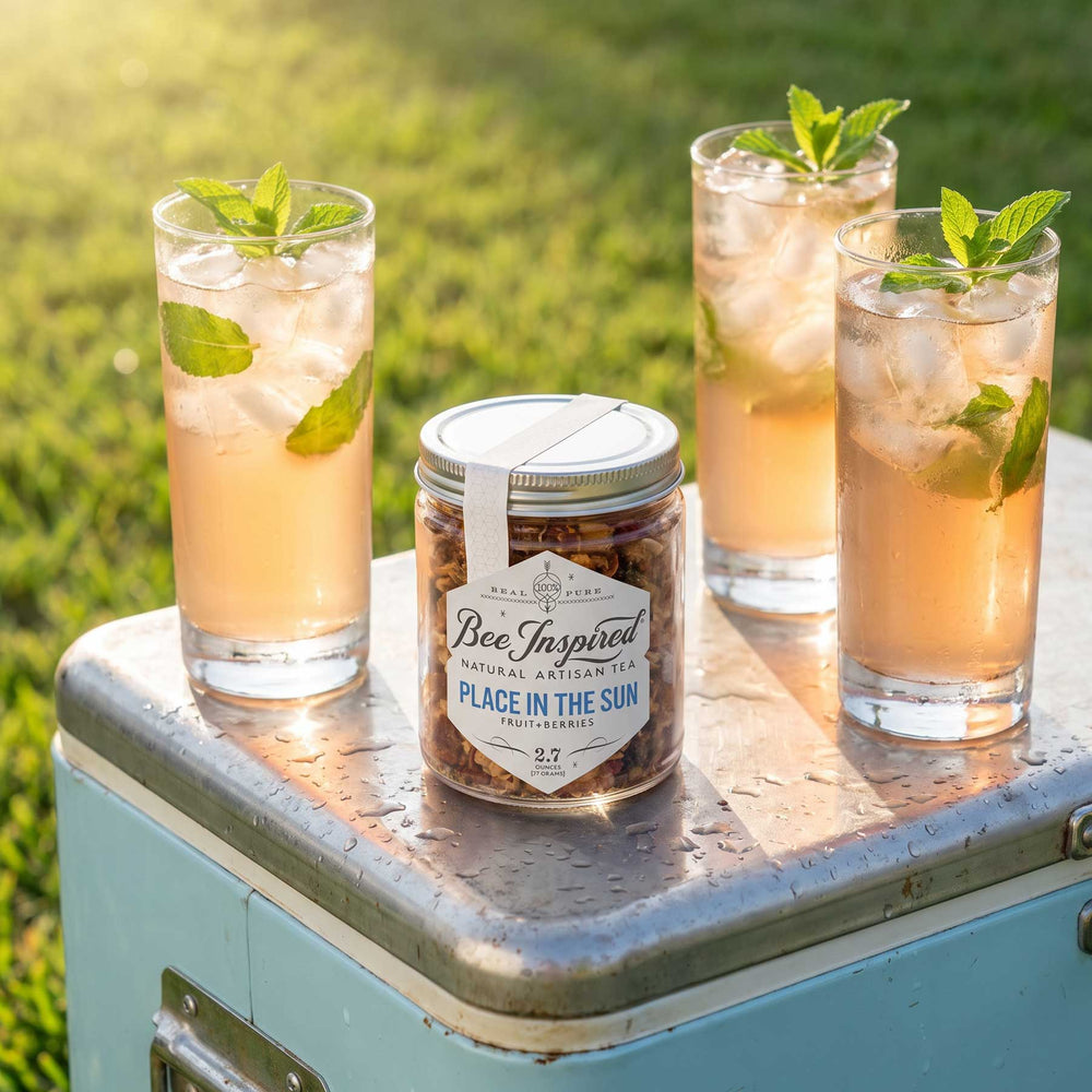 A jar of Bee Inspired natural sweeteners sits on a light blue cooler outdoors, with three glasses of iced tea garnished with mint leaves in the background on a grassy lawn.