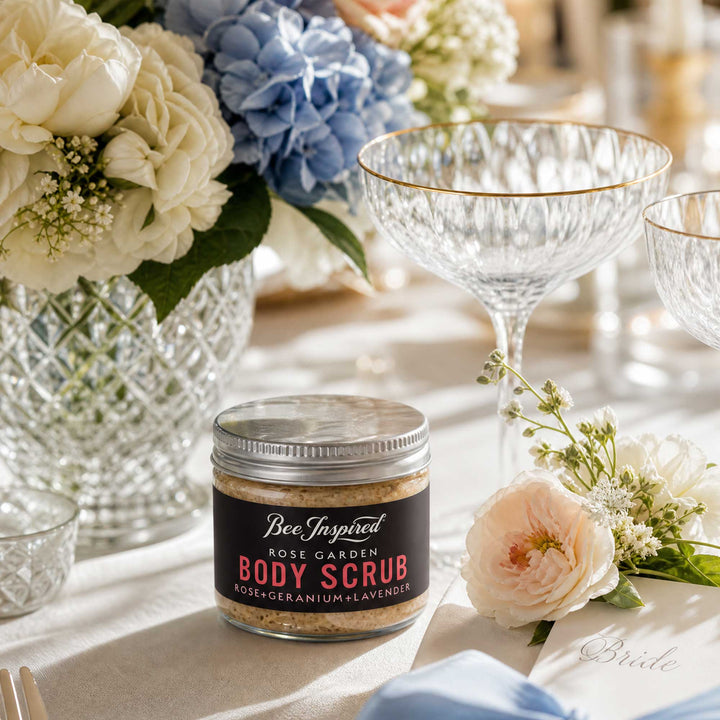 Jar of BeeInspired Rose Garden Body Scrub party favor on a table with flowers and glasses.