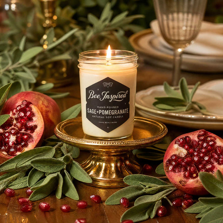 A lit candle in a clear glass jar sits on a gold pedestal, surrounded by pomegranates, sage leaves, and a warm, festive tabletop setting. The candle label reads &