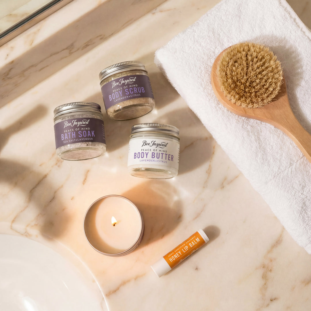 Bee Inspired skincare products including body scrub, body butter, and lip balm on a marble surface with a candle and brush.