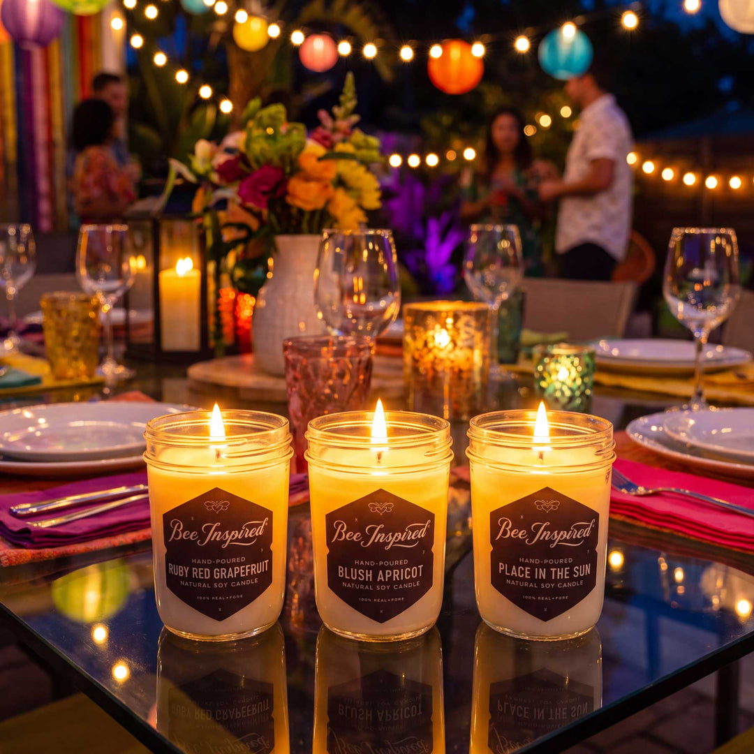 Three lit beeswax candles in clear glass jars on a decorated outdoor dining table at night, with warm string lights, flowers, and blurred people in the background. The candle labels read Bee Inspired and show scents including Ruby Red Grapefruit, Blush Apricot, and Place in the Sun.