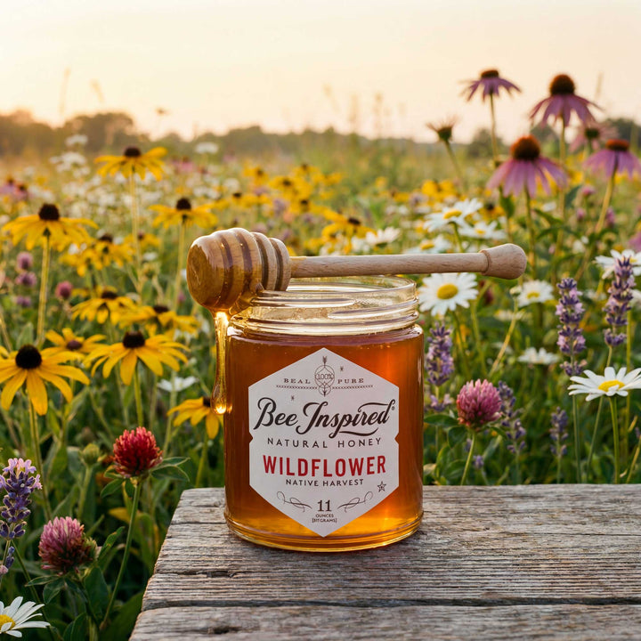 Jar of Bee Inspired Wildflower honey with a honey dipper on a wooden surface in a field of flowers.